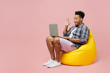 Wall Mural - Full body young man of African American ethnicity wear blue shirt sit in bag chair use work on laptop pc computer waving hand isolated on plain pastel light pink background. People lifestyle concept.