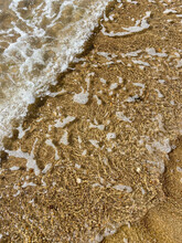 Close-up Of The Sea Coast, Clear Sea Water With Shallow Waves And A Narrow Strip Of White Foam With Bubbles Washes The Shore With A Smooth Brown Sandy Surface. High Quality Photo