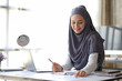 © Wasan - Attractive Muslim businesswoman working with laptop and analyzing financial statements on desk in office.