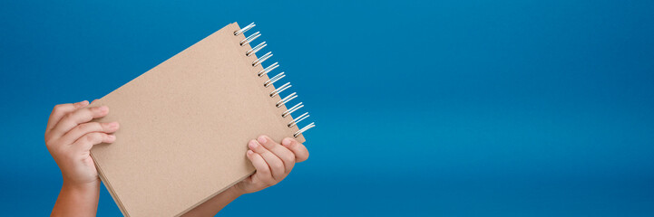 Wall Mural - Notepad in hand. Blank notepad in the hands of a child on a blue background. Hands hold a book without text. The girl is holding a book with an empty cover.