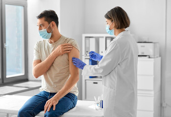 Wall Mural - health, medicine and pandemic concept - female doctor or nurse wearing protective medical mask and gloves with syringe vaccinating male patient at hospital