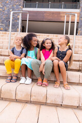 Wall Mural - Full length of multiracial smiling elementary schoolgirls sitting on steps against school building