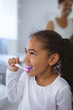 © WavebreakMediaMicro - Multiracial girl brushing teeth while looking away with father in background