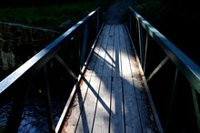 A Park Bridge With A Wooden Railing Leads Across The Stream. The Beams Are Fixed To A Steel Crossbeam That Holds In A Concrete Foundation. Road From Gravel, In Front Of The Bridge Barrier