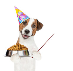 Poster - Jack russell terrier puppy wearing party cap holds  bowl of dry dog food and points away on empty space. isolated on white background