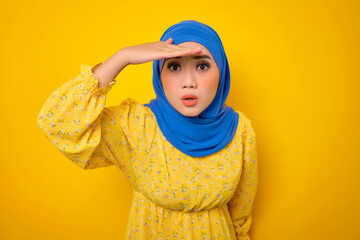 Curious young Asian woman in casual dress holding hand above eyes and peering into distance isolated on yellow background