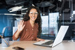 © Liubomir - Portrait of young successful business woman in modern office using laptop, Hispanic woman with curly hair smiling and looking at camera, holding hand up in greeting gesture.