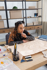 Wall Mural - Focused african american craftswoman looking at sewing pattern near equipment in workshop.