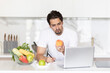 © Vitaliy - Young handsome and fit man sitting at the table in the kitchen and writing his diet plan in notebook. A lot of fruits and vegetables which on the table also included in his ration