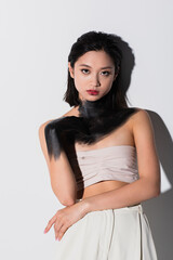 Wall Mural - young asian woman with bare shoulders and hand in black paint looking at camera on white.