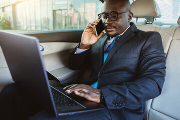 Wall Mural - Portrait of a handsome dark-skinned afro-american man in a business suit and glasses in the back seat of a luxury car is working, holding a laptop and using it