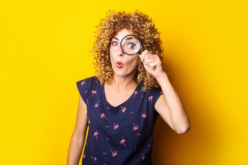 Wall Mural - surprised curly young woman looks at the camera through a magnifying glass on a yellow background.