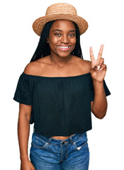 Wall Mural - Young african american woman wearing summer hat smiling with happy face winking at the camera doing victory sign. number two.