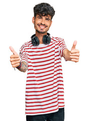 Wall Mural - Young hispanic man listening to music using headphones approving doing positive gesture with hand, thumbs up smiling and happy for success. winner gesture.