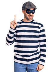 Poster - Young handsome man wearing burglar mask pointing finger up with successful idea. exited and happy. number one.