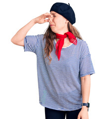 Sticker - Young beautiful blonde woman wearing french beret and striped t-shirt smelling something stinky and disgusting, intolerable smell, holding breath with fingers on nose. bad smell