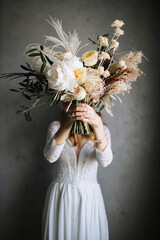 The bride in a white lace dress holds in front of her a boho-style bouquet of white and cream peonies, cereal grasses and meadow flowers.