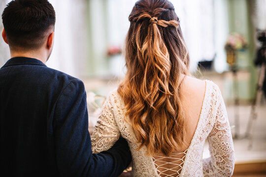 The bride and groom are behind. The bride has blond hair styled in curls and a lace-up dress with a bare back. The groom in a black jacket holds the bride by the arm.