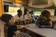 © PoppyPix - Active interracial group of four good friends on a summer road traveling, sitting inside their comfortable modern motorhome, playing the guitar, chatting and laughing. RVs interior. High quality photo