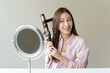© Pormezz - women hairdo makeup routine, Young woman looking at the mirror and using iron hair curling curly long hair at home.