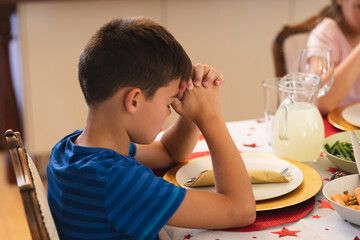 Wall Mural - Caucasian boy sitting at table for dinner with family and praying with eyes closed