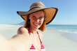Caucasian woman at beach wearing a hat and taking selfie with her smartphone