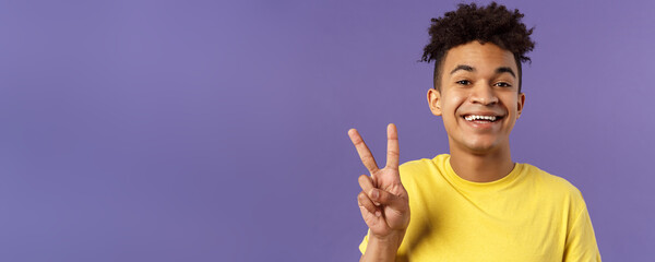 Wall Mural - Close-up portrait of handsome upbeat young teenage guy with afro hairstyle, show peace sign and smiling, wear yellow t-shirt, staying optimistic and positive, purple background