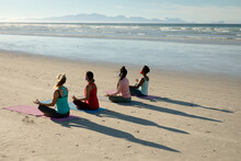 Group Of Diverse Female Friends Wearing Face Masks Practicing Yoga, Meditating At The Beach