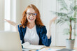 Shot of positive red haired female freelancer works remotely with paper documents, spreads hands to show her good result, sits at desktop with notepad and laptop computer, prepares course work
