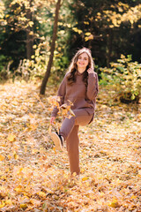 Young woman in hoodie sweater kicks up leaves in autumn park. Sunny weather. Fall season.