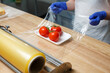 Worker wraps tray with tomatoes with cling film which he pulls from large roll standing on table.