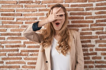 Poster - Beautiful blonde woman standing over bricks wall peeking in shock covering face and eyes with hand, looking through fingers with embarrassed expression.