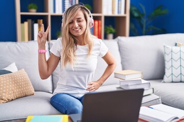 Poster - Young blonde woman studying using computer laptop at home smiling looking to the camera showing fingers doing victory sign. number two.