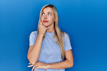 Poster - Beautiful blonde woman wearing casual t shirt over blue background with hand on chin thinking about question, pensive expression. smiling with thoughtful face. doubt concept.