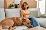 Fototapeta Zwierzęta - Young caucasian woman using laptop sitting on sofa with dog at home