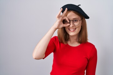 Poster - Young redhead woman standing wearing glasses and beret doing ok gesture with hand smiling, eye looking through fingers with happy face.