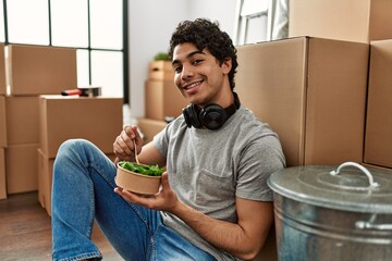 Canvas Print - Young hispanic man eating salad sitting on the floor at new home.