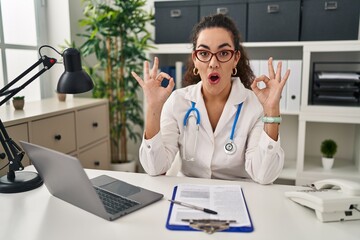 Sticker - Young hispanic woman wearing doctor uniform and stethoscope looking surprised and shocked doing ok approval symbol with fingers. crazy expression