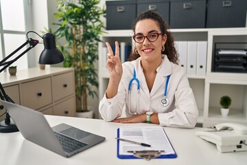 Sticker - Young hispanic woman wearing doctor uniform and stethoscope smiling with happy face winking at the camera doing victory sign with fingers. number two.