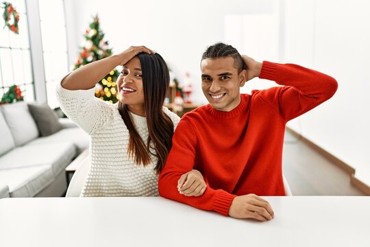 Young latin couple sitting on the table by christmas tree smiling confident touching hair with hand up gesture, posing attractive and fashionable