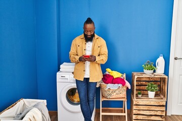 Poster - Young african american man using smartphone waiting for washing machine at laundry room