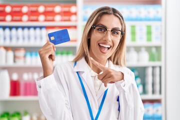 Poster - Young blonde woman working at pharmacy drugstore holding credit card smiling happy pointing with hand and finger