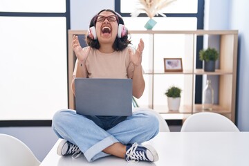 Canvas Print - Young hispanic woman using laptop sitting on the table wearing headphones celebrating mad and crazy for success with arms raised and closed eyes screaming excited. winner concept