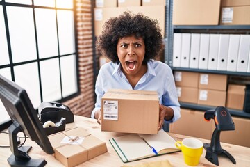 Wall Mural - Black woman with curly hair working at small business ecommerce holding box angry and mad screaming frustrated and furious, shouting with anger looking up.
