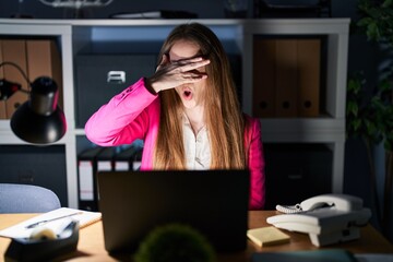 Canvas Print - Young caucasian woman working at the office at night peeking in shock covering face and eyes with hand, looking through fingers with embarrassed expression.