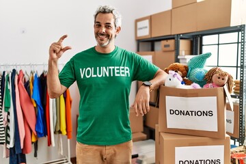 Wall Mural - Middle age hispanic man wearing volunteer t shirt at donations stand smiling and confident gesturing with hand doing small size sign with fingers looking and the camera. measure concept.