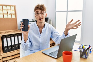 Wall Mural - Young caucasian businessman working using laptop and showing smartphone celebrating achievement with happy smile and winner expression with raised hand