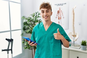 Canvas Print - Young caucasian physio man using touchpad at the clinic smiling happy and positive, thumb up doing excellent and approval sign