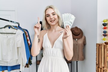 Wall Mural - Young caucasian woman holding dollars at retail shop smiling with an idea or question pointing finger with happy face, number one