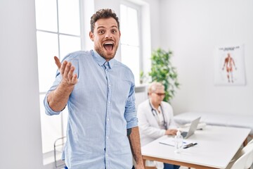 Sticker - Hispanic man at doctor clinic celebrating achievement with happy smile and winner expression with raised hand